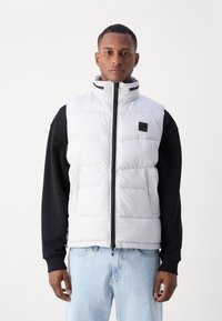 Gilet matelassé blanc avec col haut, zip frontal et poches latérales. Les accents noirs comprennent le logo et des manches contrastantes. Texture douce et matelassée.