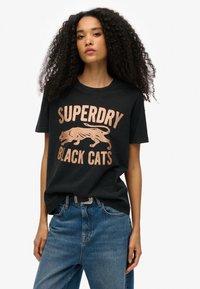 Camiseta negra con el texto "SUPERDRY BLACK CATS" en color rosa dorado metálico, que presenta un gráfico de un gato corriendo. Ajuste casual, material de algodón.