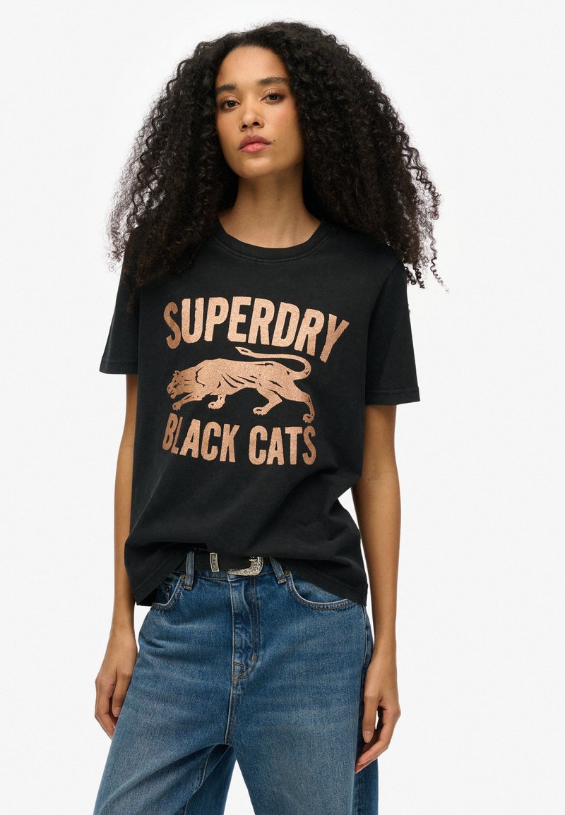 Camiseta negra con el texto "SUPERDRY BLACK CATS" en color rosa dorado metálico, que presenta un gráfico de un gato corriendo. Ajuste casual, material de algodón.