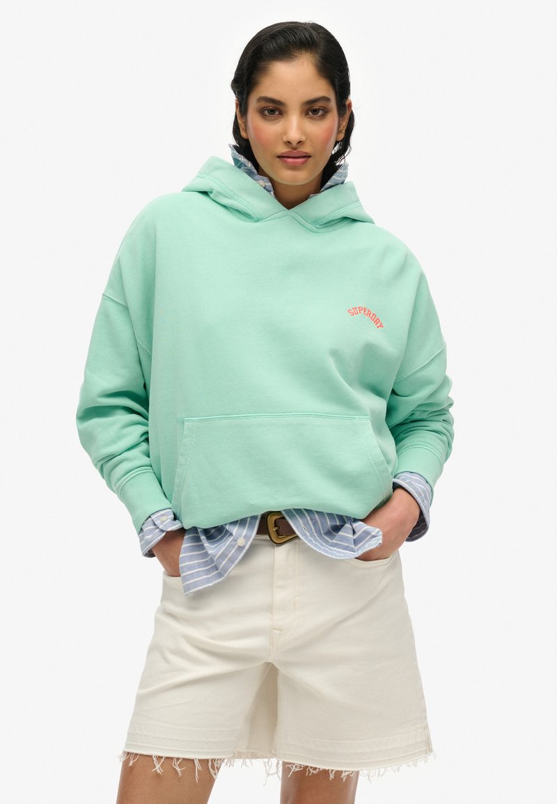Muntgroene hoodie met een voorkantzak en een rood "Superdry" logo. Draag het over een lichtblauw gestreept shirt en in combinatie met witte shorts.