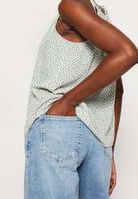 Débardeur vert à motif floral avec des fleurs blanches, porté avec un jean taille haute bleu clair. Le tissu semble léger avec une coupe décontractée.