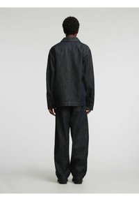 Tumša džinsa jaka un bikses ar vaļīgu piegriezumu, taisnu griezumu un redzamām šuvēm. Auduma tekstūra ir gluda ar vieglu spīdumu.