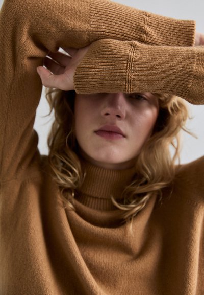 Barna kötött pulóver magas nyakkal és bordázott mandzsettákkal; az anyag puhanak és texturált hatású, laza szabással és felgöngyölt gallérral rendelkezik.