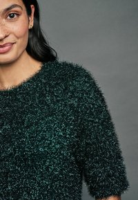 Maglione fuzzy nero e verde petrolio con una consistenza morbida, maniche corte e un ampio scollo, caratterizzato da un dettaglio in tessuto denso e peloso.