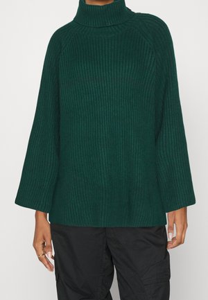 Pull à col roulé côtelé vert foncé avec manches longues et larges, porté avec un pantalon cargo noir.
