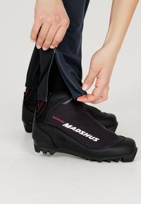 Svarta nordiska skidpjäxor med en texturerad yta och röda detaljer, kombinerade med marinblå byxor som har en zippered nederdel för enkel justering.