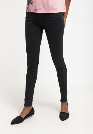 Person trägt schwarze enge Hosen und schwarze spitze Ballerinas, steht mit einer Hand in der Tasche vor einem weißen Hintergrund.