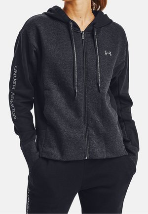 Kobieta ubrana w ciemnoszarą i czarną bluzę Under Armour na zamek z logo na piersi i tekstem na rękawie, ręce w kieszeniach.
