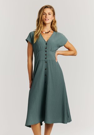 Vrouw die een teal midi-jurk met knopen voor en korte mouwen draagt, staande met één hand op haar heup tegen een effen lichte achtergrond.