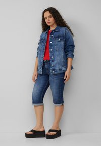 Denim jack in middelblauw, met knopenlijst, twee borstzakken en opgerolde mouwen. Gecombineerd met een rode top en denim capri-broeken. Zwarte plateausandalen.