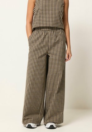 Vrouw draagt beige en zwart geruite wijdvallende broek en bijpassende mouwloze top met witte sneakers, staand met handen in de zakken.