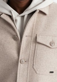 Beige Jacke aus Wollmischung mit Knopfverschluss, Brusttasche und weicher Textur; darüber ein hellgrauer Hoodie mit strukturierter Stoffoberfläche.