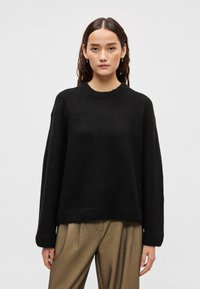 Μαύρο πλεκτό πουλόβερ με στρογγυλή λαιμόκοψη, κατεβασμένους ώμους και ριμπ μανσέτες. Φορεμένο πάνω από ανοιχτό καφέ ψηλόμεσα παντελόνια.