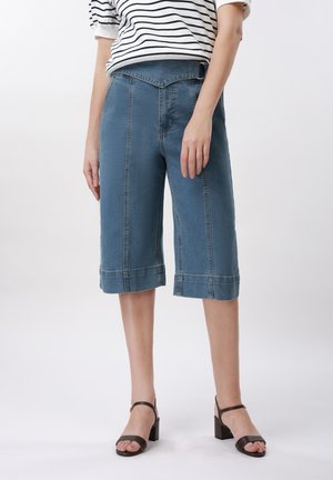 Femme portant un short en jean mi-long avec des coutures à l'avant, un haut rayé blanc et noir, et des sandales noires à talons blocs, debout.