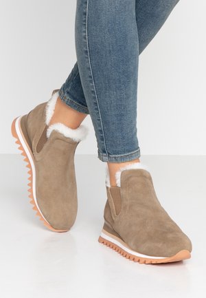 Fødder iført lysebrune ruskinds slip-on-støvler med hvid pelsforing og ribbet lysebrun sål, kombineret med blå denim skinny jeans.