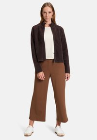 Giacca in pile marrone scuro, corta, con colletto alto e chiusura a zip. Indossata sopra una camicia bianca e abbinata a pantaloni larghi marroni e sneakers bianche.