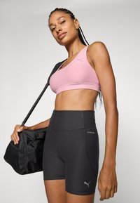 Helle rosa Sport-BH und enganliegende schwarze Shorts mit hoher Taille. Beide haben eine glatte Textur; die Shorts sind mit einem Puma-Logo und dem Text "CLOUDSPUN" versehen.