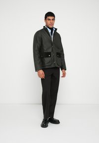 Ανθεκτικό στο νερό μπουφάν ελιάς με κούμπωμα με κουμπιά και δύο μπροστινές τσέπες με μαύρες βελούδινες λεπτομέρειες. Φορεμένο πάνω από σκουρόχρωμο παντελόνι και παπούτσια.