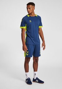 Marinblå idrottströja och shorts med limegröna accenter. Tröjan har korta ärmar medan shortsen har ett chevronmönster. Vita strumpor och skor fulländar looken.