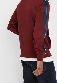 Persoon in een bordeauxrood T-shirt met lange mouwen met marineblauwe en witte strepen op de mouwen, donkerblauwe jeans en een wit shirt eronder.