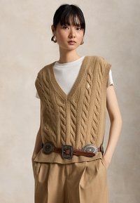 Tânără purtând un vestă bej tricotată cu model torsadat peste un tricou alb, accesorizată cu o curea lată din piele cu conchos metalici, pantaloni bej.
