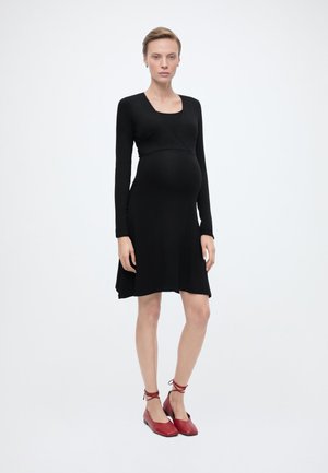 Terhes nő áll, fekete hosszú ujjú ruhát és piros bokapántos balerinacipőt visel, egyszínű fehér háttér előtt.