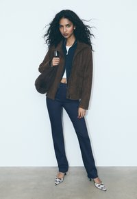 Hnědá semišová bunda, tmavě modré kalhoty, bílý krátký top a vzorované podpatky. Modelka pózuje s hnědou taškou na jednobarevném pozadí.