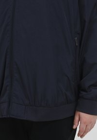 Jachetă ușoară de culoare albastru închis, cu un fermoar frontal, manșete elastice și un tiv elastic, având un buzunar lateral cu fermoar și o textură netedă.