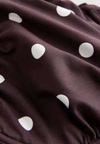 Tissu marron foncé avec des pois blancs irréguliers, montrant des plis doux et une texture.