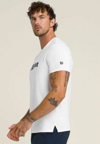 Hombre con cabello rizado lleva una camisa de manga corta blanca con texto negro y tatuajes visibles en los brazos, contra un fondo plano.