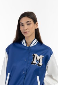 Giacca varsity in satin blu con maniche bianche, caratterizzata da una grande lettera "M" ricamata sul petto e colletto e polsi a righe ribeccati.