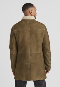 Olivenfarbener Wildledermantel mit Schaffellkragen, langen Ärmeln und kontrastierenden Nähten. Verfügt über ein Rückenlabel und eine taillierte Passform.