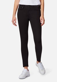 Jeans ajustados negros de mezclilla con una textura suave. Cinco bolsillos y pasadores para el cinturón; combinados con zapatillas blancas. Corte slim a lo largo de las piernas.