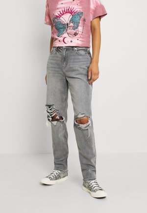 Osoba nosząca szare, podarte dżinsy, różowy t-shirt z niebieskim motylem i grafiką słońca oraz białe trampki z wzorem skóry węża.