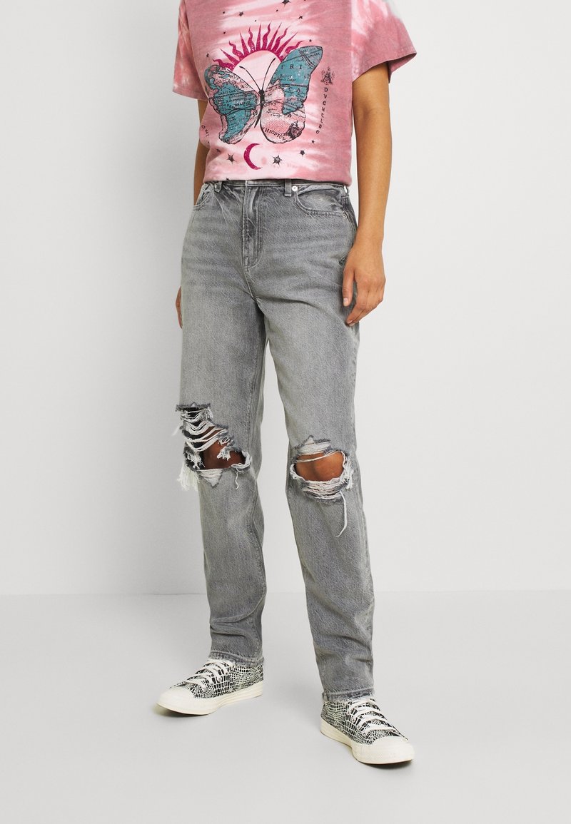 Personne portant un jean gris déchiré, un t-shirt rose avec un papillon bleu et un motif de soleil, et des baskets blanches à motif peau de serpent.