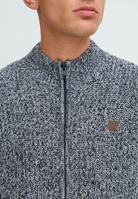 Megztas zip-up megztinis tamsiai mėlynos ir grietinėlės spalvos, su aukšta apykakle, tekstūrine raštu ir maža ruda odine logotipo etikete ant krūtinės.