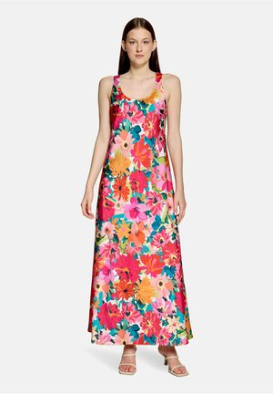 Kobieta stoi, ubrana w bezrękawnikową sukienkę do kostek z żywym, wielokolorowym kwiatowym wzorem oraz beżowe sandały na obcasie z odkrytymi palcami.