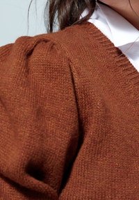 Pull en tricot marron avec détail d'épaule bouffante porté par-dessus une chemise blanche à col, montrant de près la texture et le tissu.
