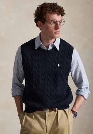 Jeune homme aux cheveux bouclés portant des lunettes rondes, un gilet en maille torsadée bleu par-dessus une chemise rayée bleue, un pantalon beige et une montre-bracelet.