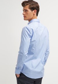 Cămașă pentru bărbați, de culoare albastru deschis, cu mâneci lungi, guler cu butoni, croială ajustată și textură netedă. Include un yoke pe spate și manșete ajustabile.