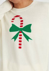 Maglione bianco con una grafica di bastoncini di zucchero a strisce rosse e bianche con un fiocco verde. Il materiale sembra morbido e ha una vestibilità ampia.