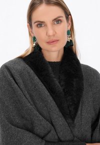 Wrap din lână gri cu guler din blană falsă neagră. Modelul poartă cercei cu ciucuri verzi și aurii, punând în evidență designul elegant al articolului.
