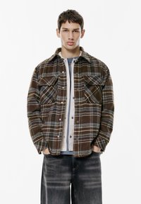 Camicia di flanella a quadri marrone con due tasche sul petto, bottoni a pressione e una maglietta beige a girocollo sotto. Jeans grigio scuro a gamba larga.