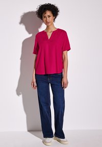 Top fucsia a maniche corte con scollatura a V, abbinato a jeans dritti blu scuro e sneakers chiare, indossato da un modello su uno sfondo neutro.