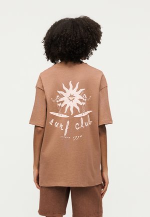 Άτομο με σγουρά μαλλιά που φοράει καφέ μπλουζάκι και σορτς, στο πίσω μέρος του μπλουζακιού υπάρχουν ήλιος, δύο σερφ και το κείμενο "surf club since 1990".