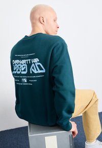 Felpa color teal con stampa grafica sul retro; presenta polsini a costine e una vestibilità rilassata. Modello seduto su una superficie metallica, indossando pantaloni color cammello.