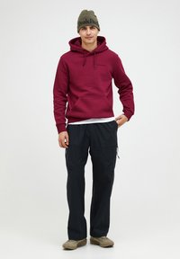 Felpa maroon con cappuccio e coulisse, tasca frontale; pantaloni cargo neri con tasche zip; sneakers beige; berretto olivastro con logo del brand.