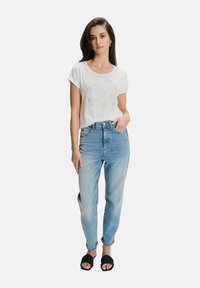 Kobieta stoi z ręką w kieszeni, ubrana w białą koszulkę z podwiniętymi rękawami, jasnoniebieskie jeansy z wysokim stanem, czarne sandały i kolczyki w kształcie kół.