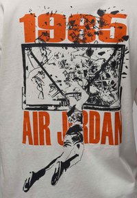 Weißes T-Shirt mit grafischem Design eines Spielers, der durch einen zerbrochenen Basketballkorb dunkt. Text: "1985" in Orange oben und "AIR JORDAN" unten.