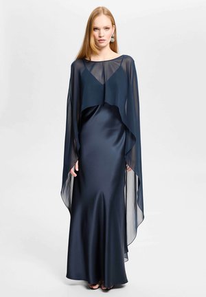Frau trägt ein bodenlanges marineblaues Satinkleid mit transparentem marineblauem Cape und silbernen Statement-Ohrringen, steht vor einem schlichten weißen Hintergrund.